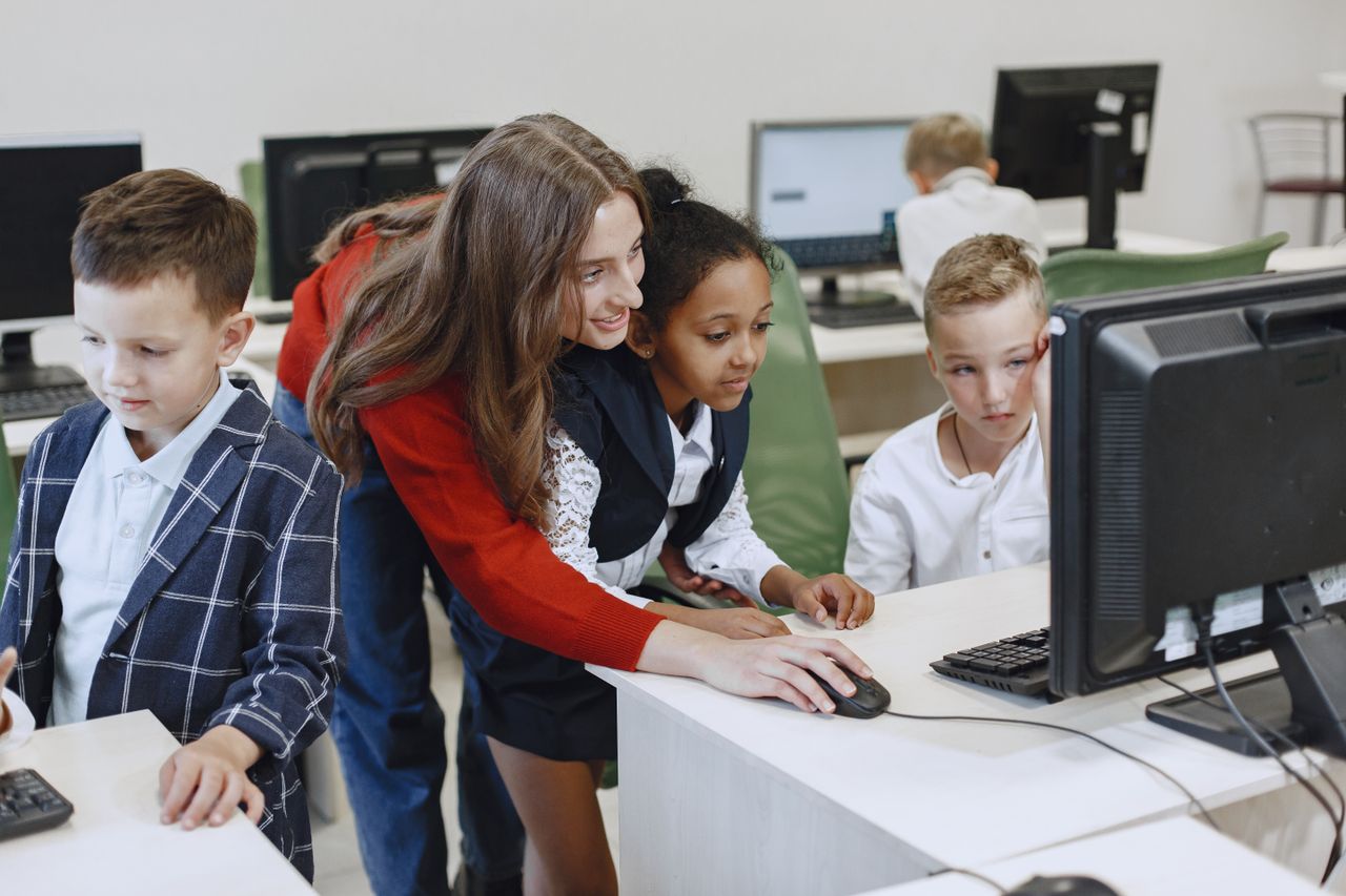 Home Teacher engaging with diverse children in a technology lesson using computers in a modern classroom.